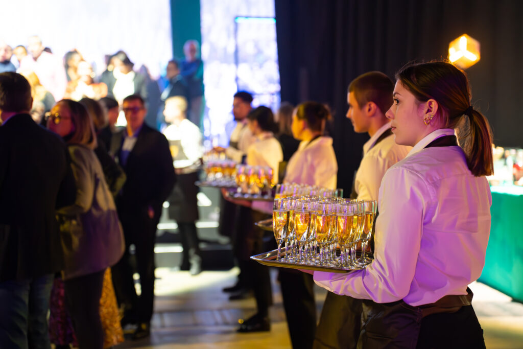 Serveuse en chemise blanche portant plateau de flûtes de champagne lors d’un cocktail d’entreprise devant une foule.