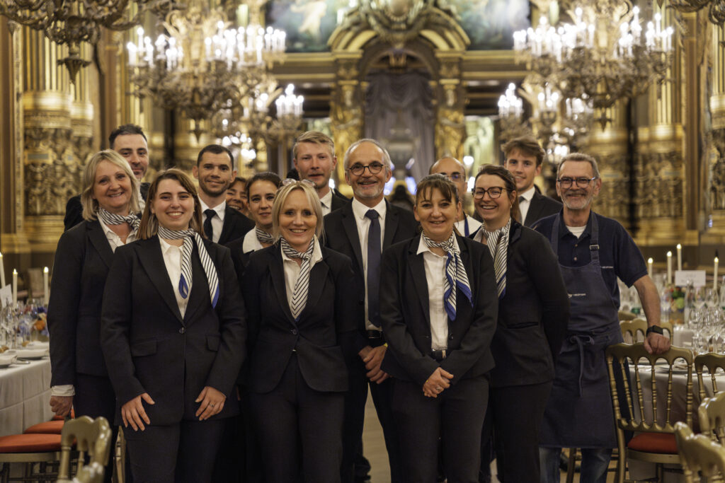 Groupe de serveurs en uniforme noir et foulards rayés, dans une salle de banquet dorée et ornée de lustres.