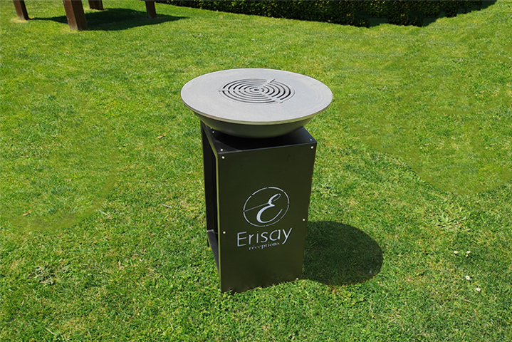 Cendrier d'extérieur noir sur socle métallique, plateau rond perforé; logo Erisay sur pelouse d'un parc.