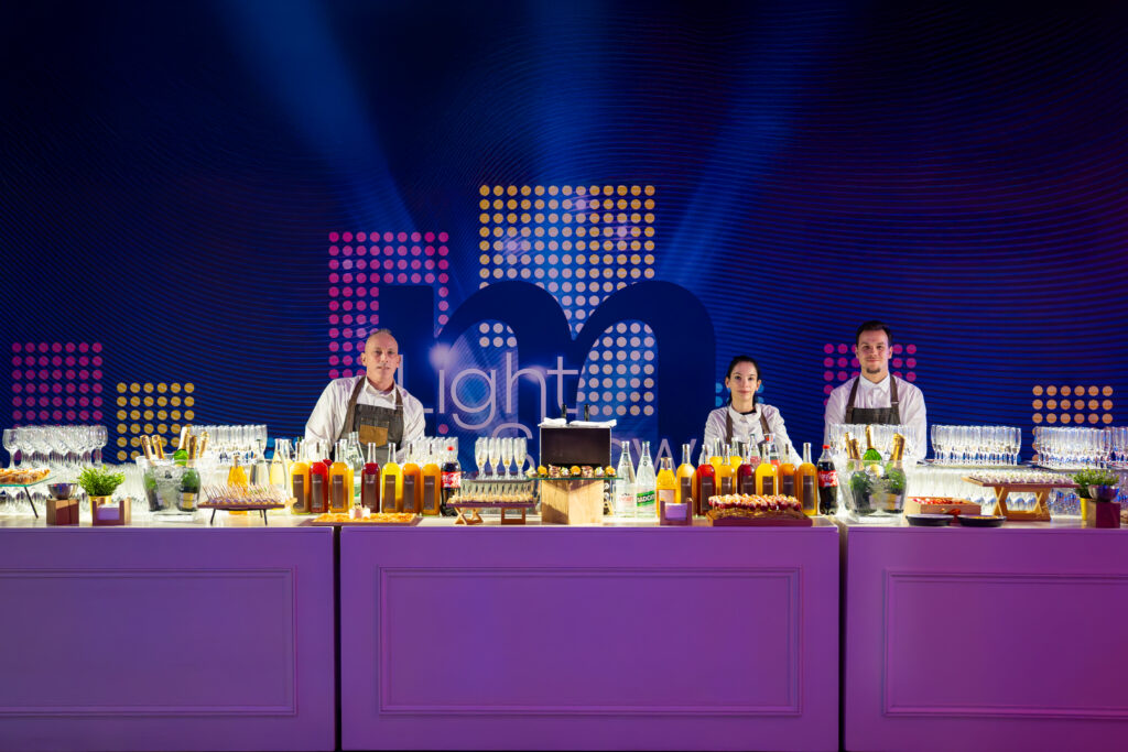 Quatre barmans en tabliers blancs derrière un bar violet, bouteilles et verres, décor lumineux à pois.