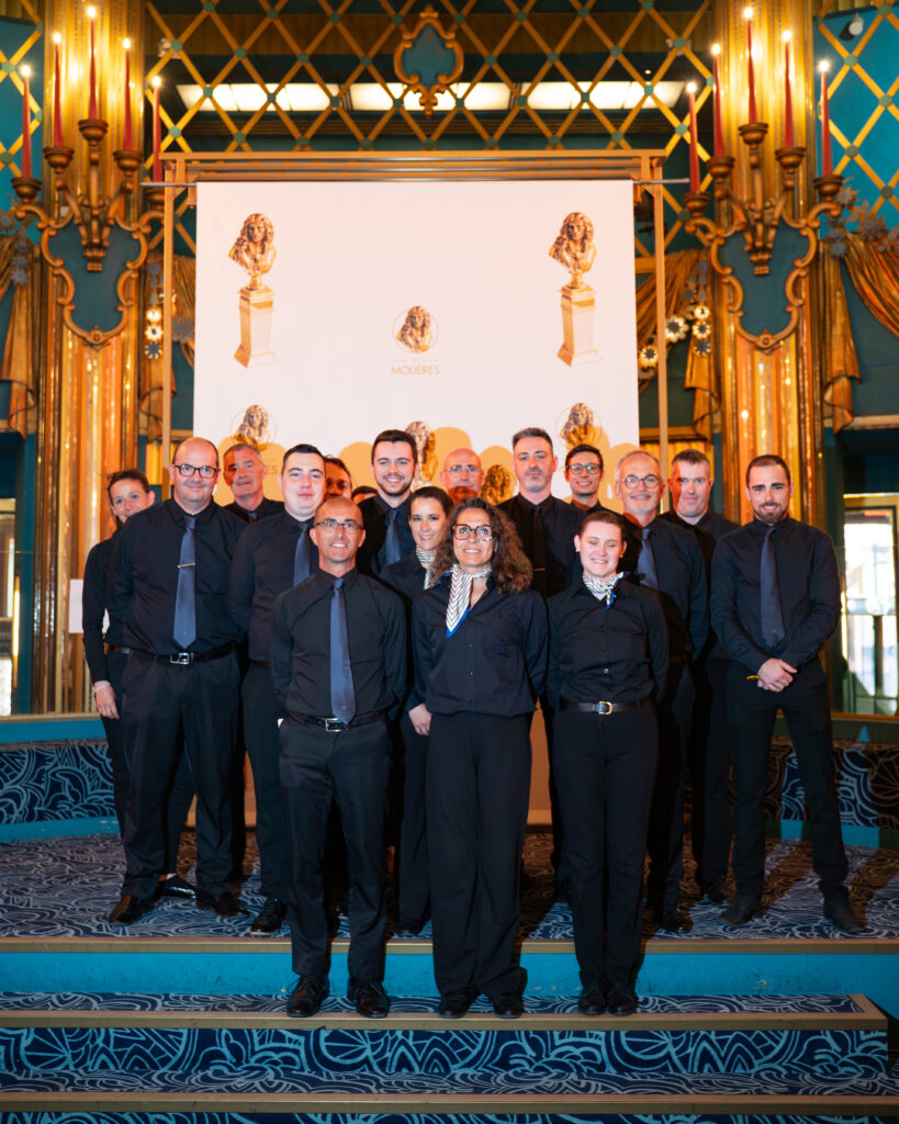 Photo de groupe de personnes en noir sur scène, décor doré et bleu lors d’un événement.