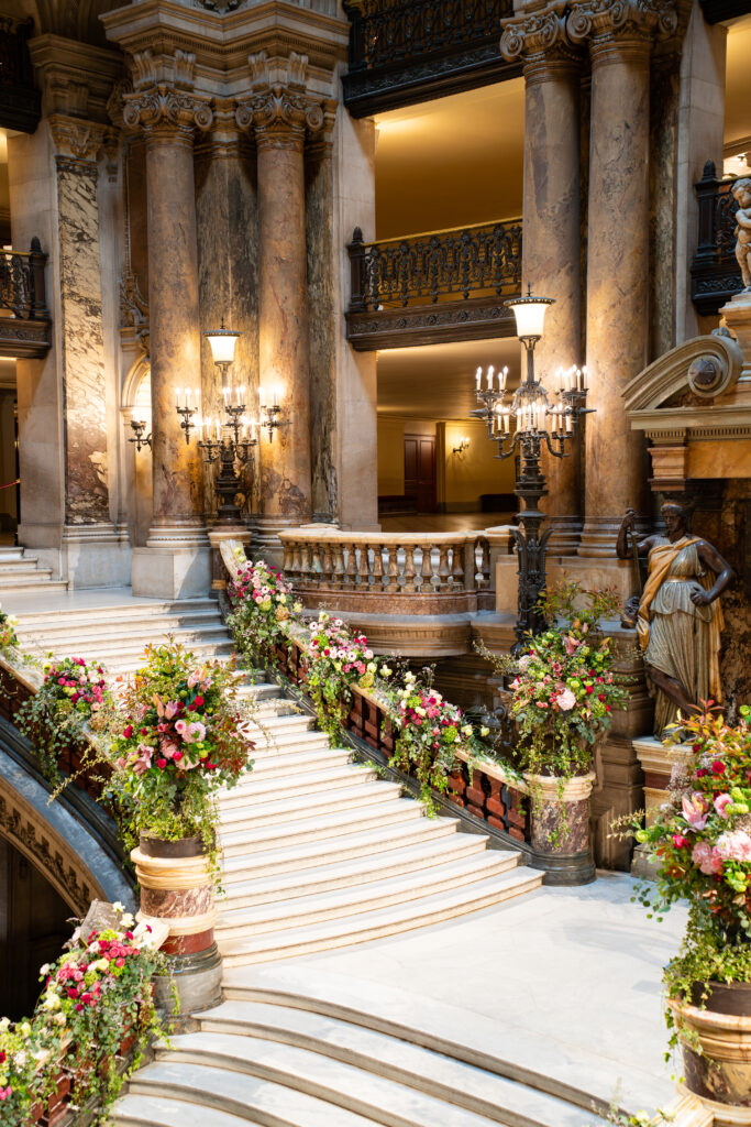 Hall majestueux en marbre avec colonnes ioniques, balustrade ornée, lustres et fleurs sur les escaliers.