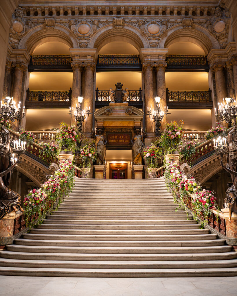 Grand escalier majestueux dans un hall théâtral, colonnes de marbre, balustrades sculptées et fleurs colorées.
