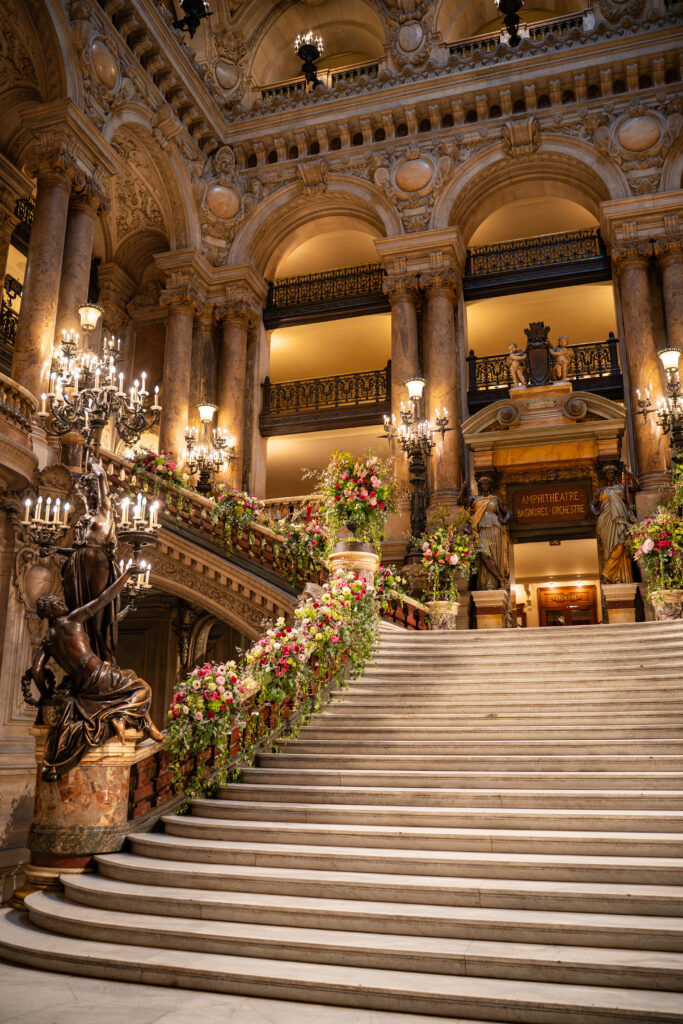 Grand escalier en marbre décoré de fleurs, dans un intérieur d’opéra richement sculpté et éclairé par des lustres.