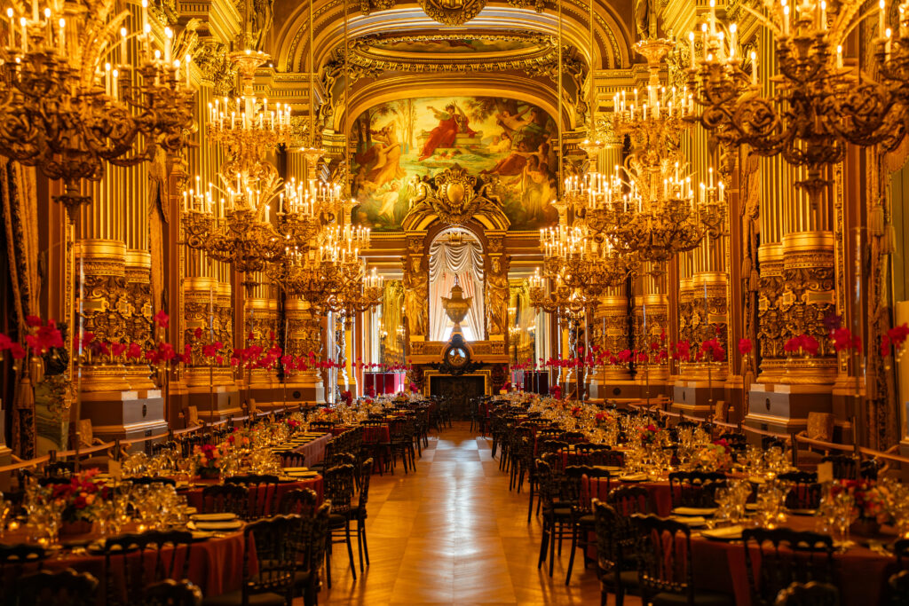 Salle de banquet dorée et opulente, lustres scintillants et fresque centrale, tables dressées pour un dîner.