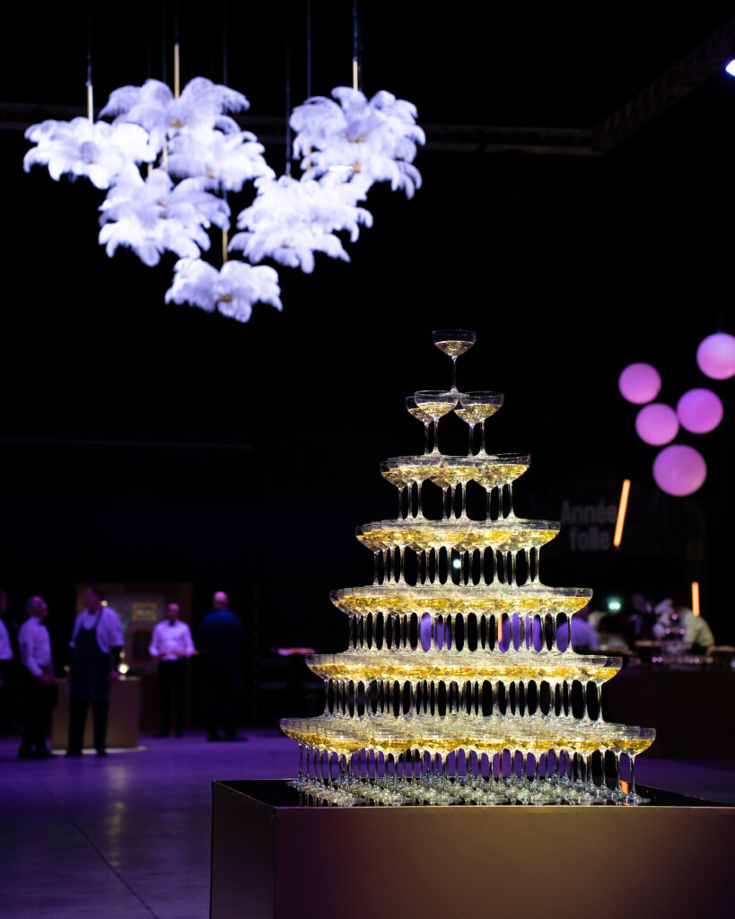 Tour de verres à champagne illuminés, décor nuages blancs suspendus et lumières violettes en arrière-plan.