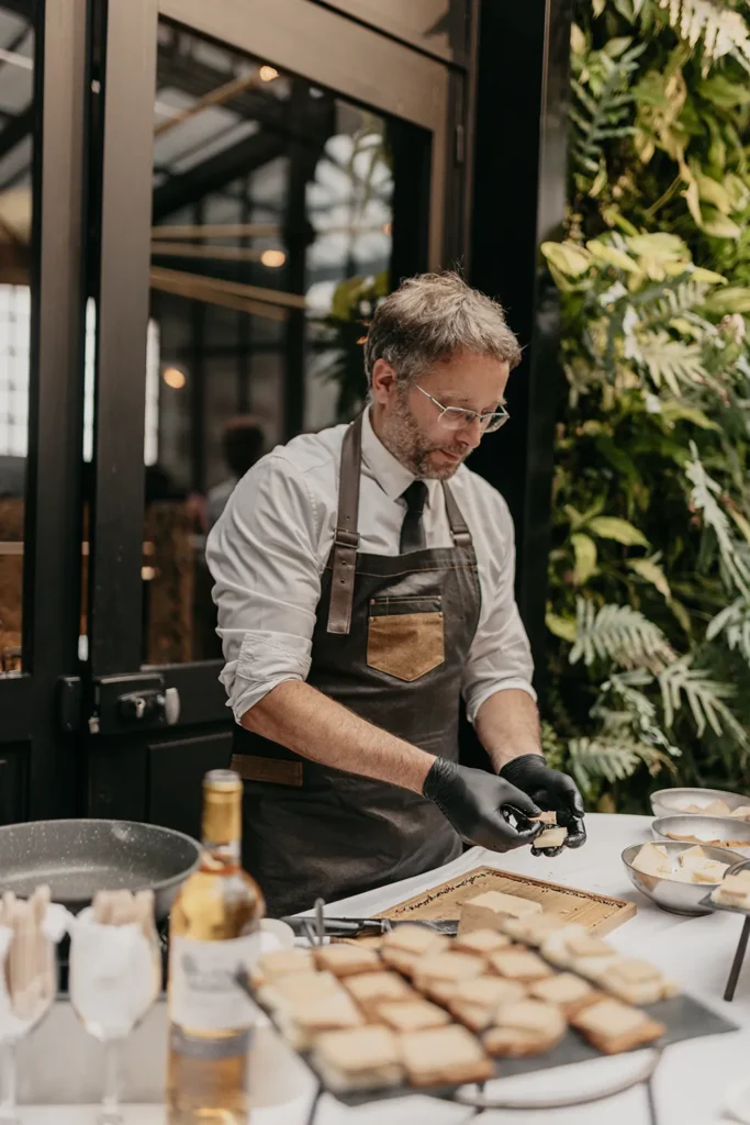 Chef en tablier sombre et gants noirs découpe des toasts sur une planche, près d'un buffet de pains.