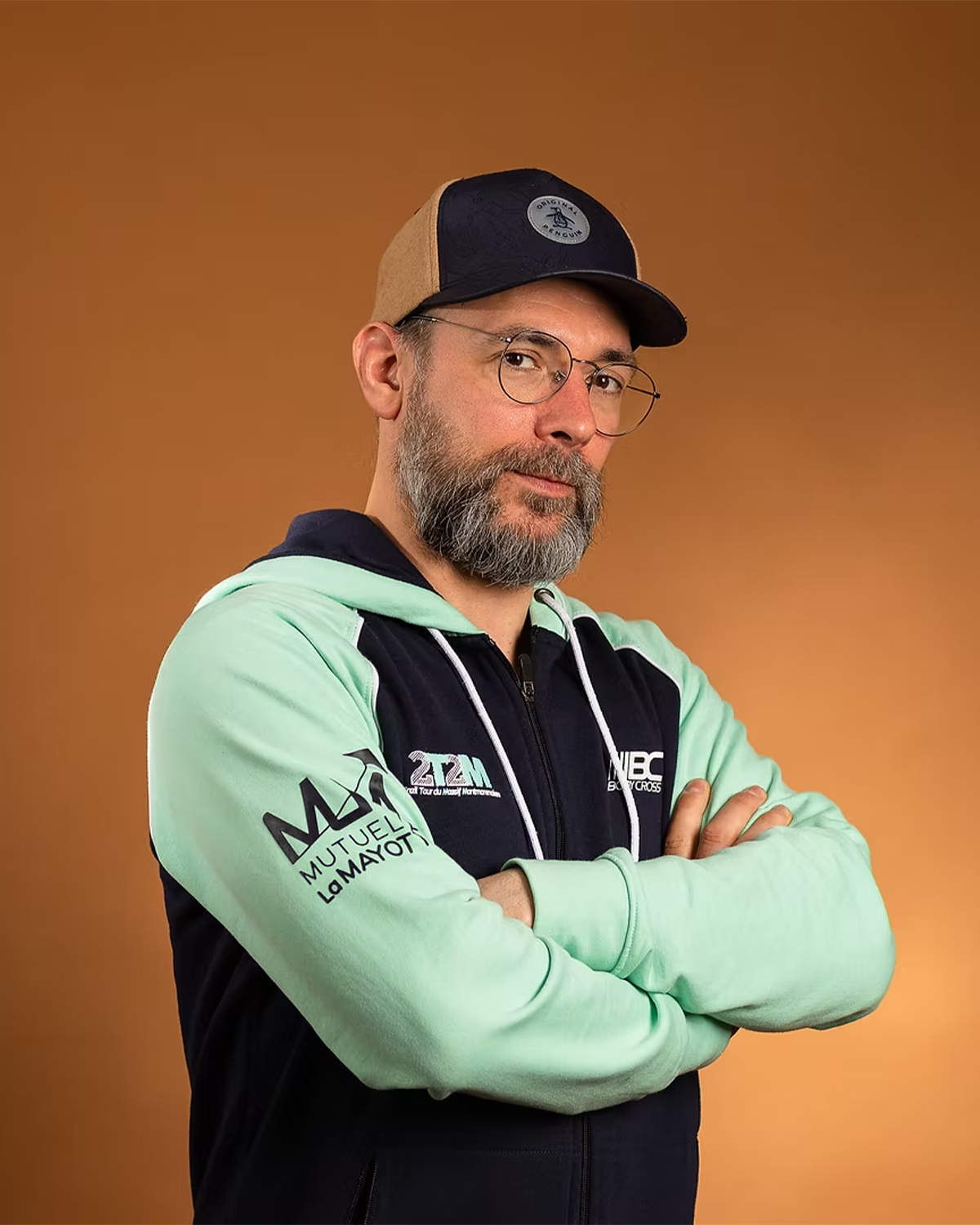 Homme portant casquette bicolore, lunettes rondes, hoodie vert menthe et marine, bras croisés sur fond orange.