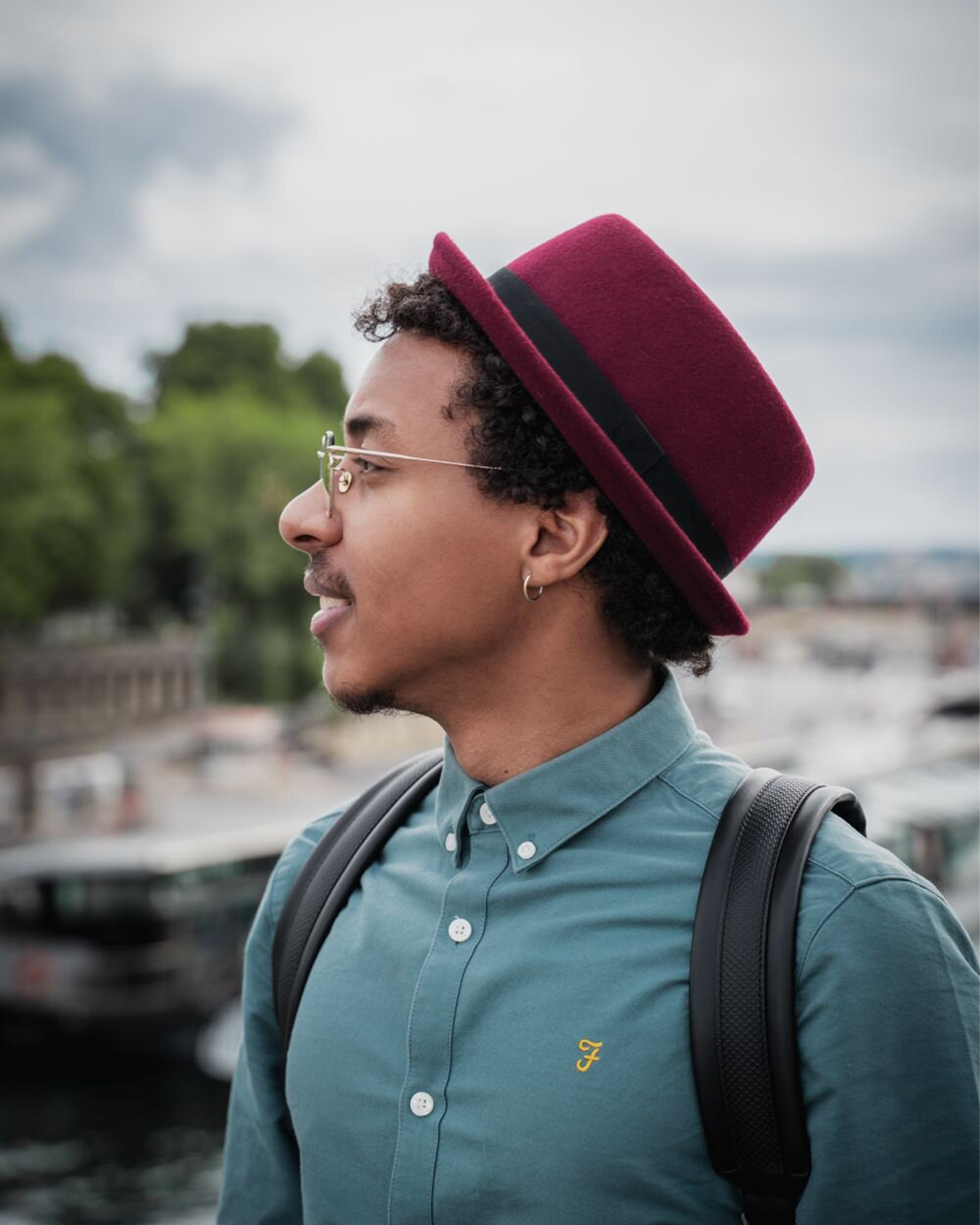 Portrait en profil d'un homme portant un chapeau bordeaux, lunettes et sac à dos, en extérieur.