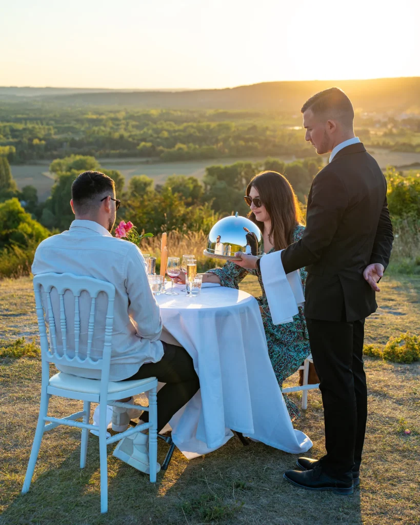 Groupe de personnes lors d'un repas en plein air au coucher du soleil; un serveur en smoking présente une cloche argentée.