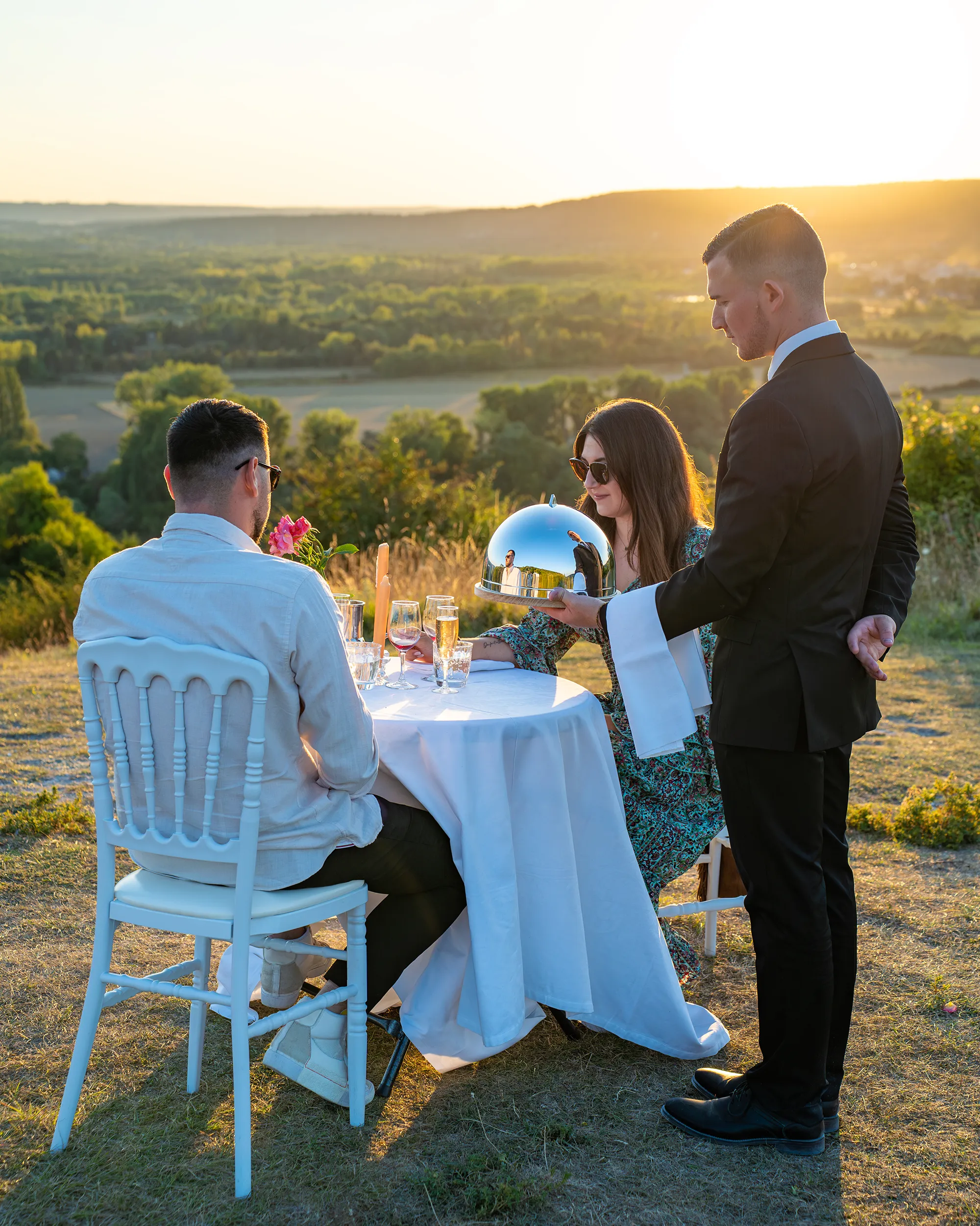 Groupe de personnes lors d'un repas en plein air au coucher du soleil; un serveur en smoking présente une cloche argentée.