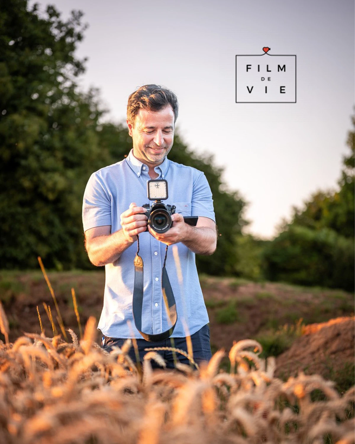 Homme souriant tenant un appareil photo avec torche LED dans un champ; logo Film de Vie au coin supérieur droit.