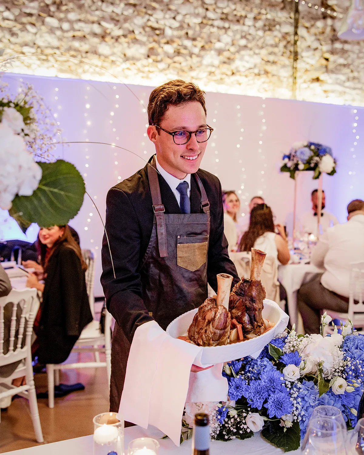 Serveur en tablier présente un plat de viande rôtie lors d'un banquet, décoré de fleurs bleues et guirlandes lumineuses.
