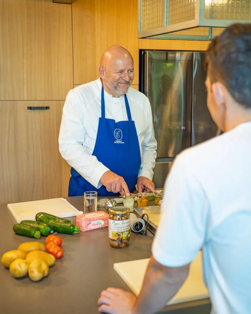 Chef souriant en tablier bleu découpe des légumes sur le comptoir, client en face dans une cuisine professionnelle.