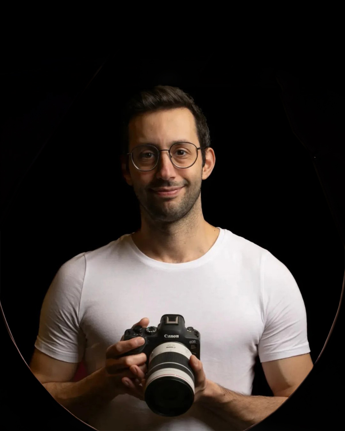 Homme portant lunettes tient un appareil photo Canon avec grand objectif, chemise blanche, fond noir.