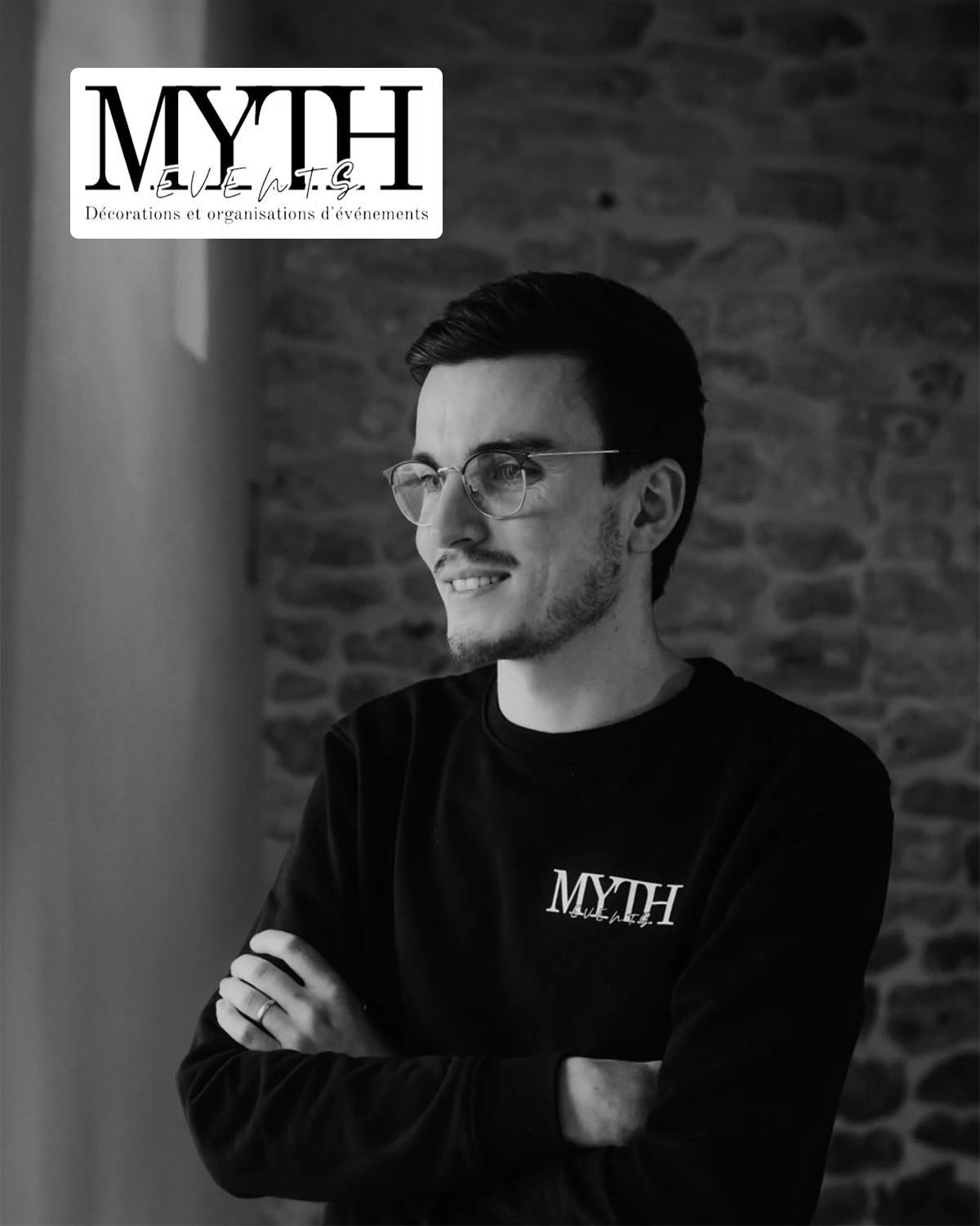 Portrait noir et blanc d’un homme souriant, lunettes et pull noir MYTH, devant mur de briques; logo MYTH Events en haut.