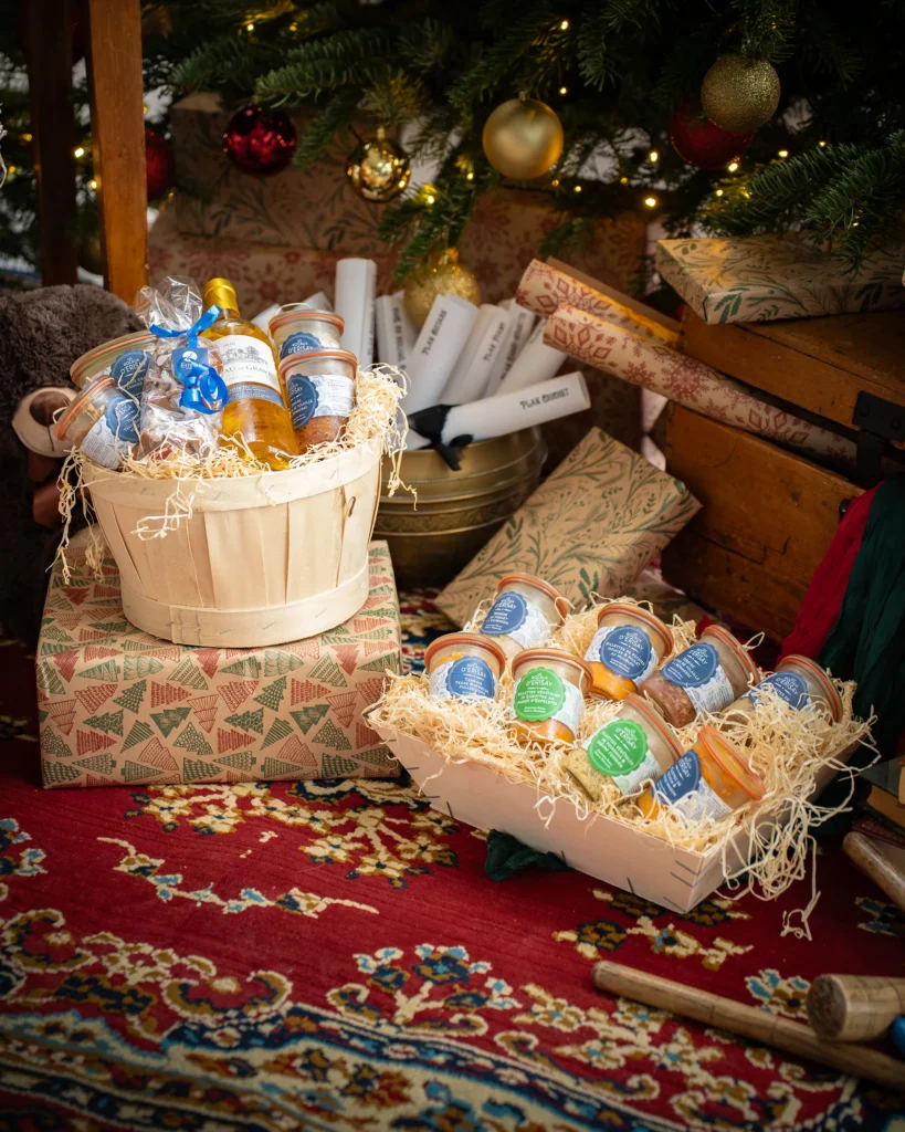 Coffrets cadeaux et bocaux de confiture dans un seau en bois, sous sapin décoré et guirlande lumineuse.