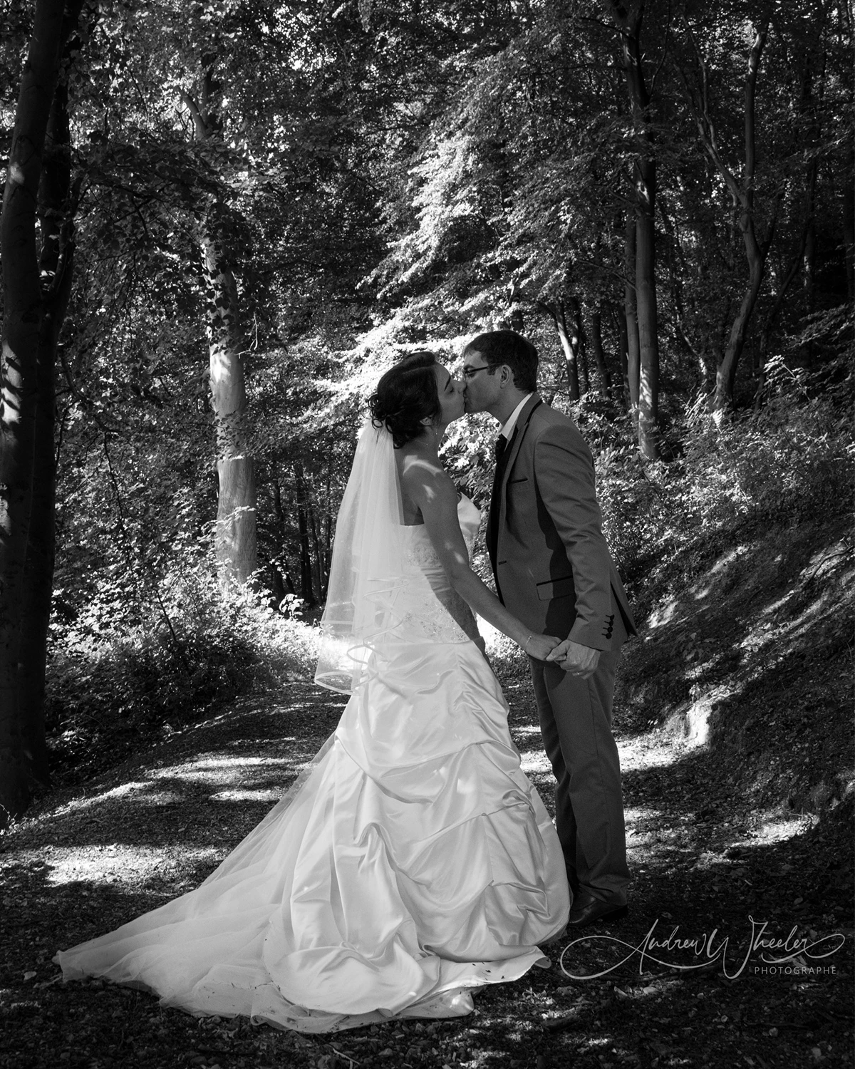 Deux mariés s'embrassent sur un chemin forestier en noir et blanc; mariée en robe blanche et voile, marié en costume.