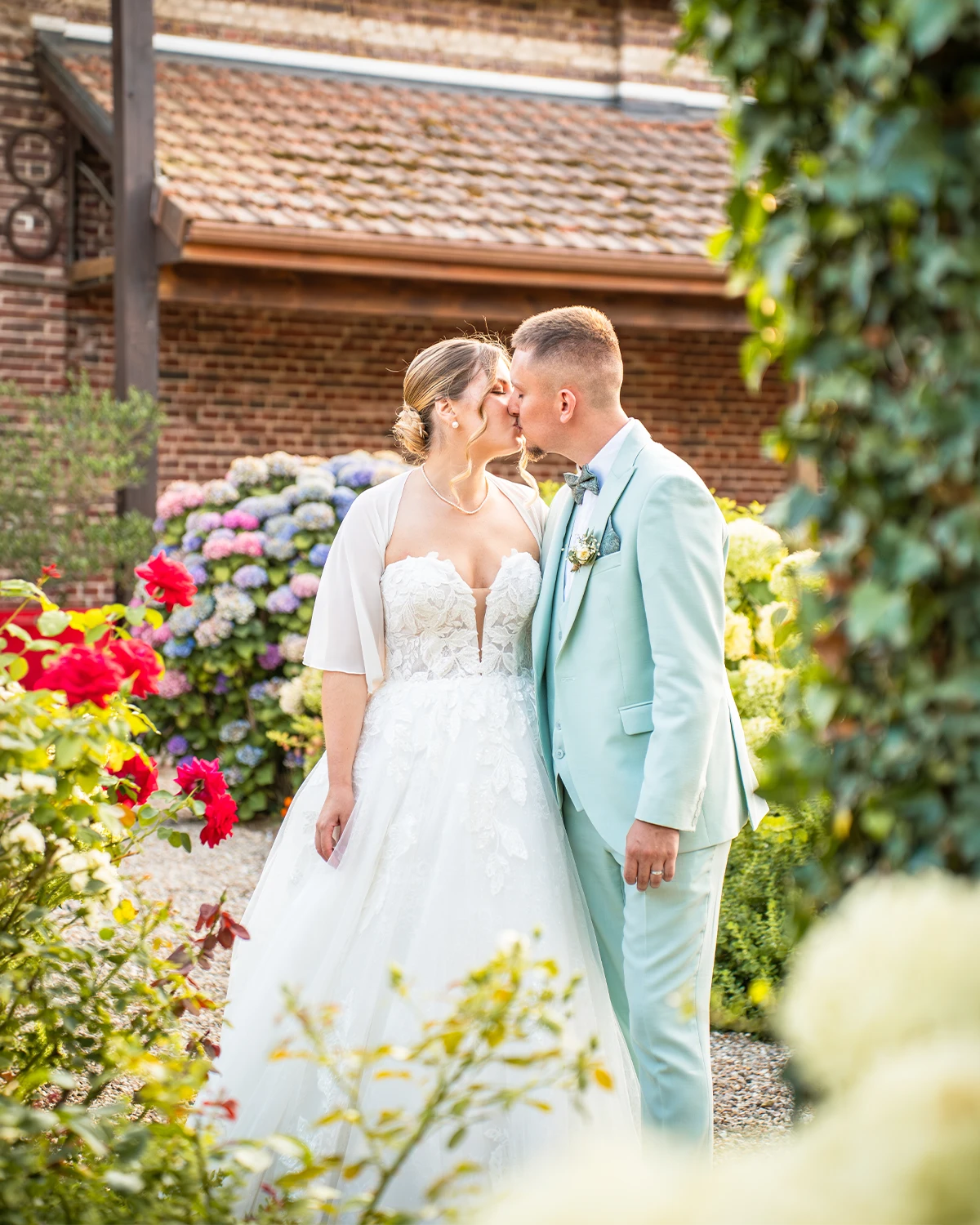 Couple de mariés s’embrassant dans un jardin fleuri, la mariée en robe blanche et le marié en costume bleu clair.