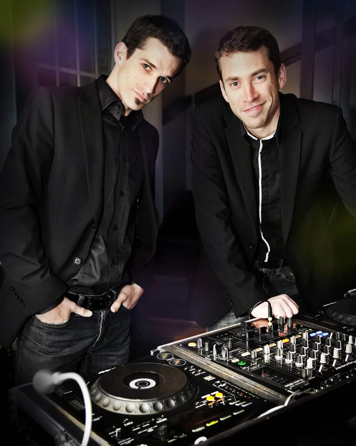 Deux DJs en vestes noires près d'une table de mixage avec platines et boutons colorés.