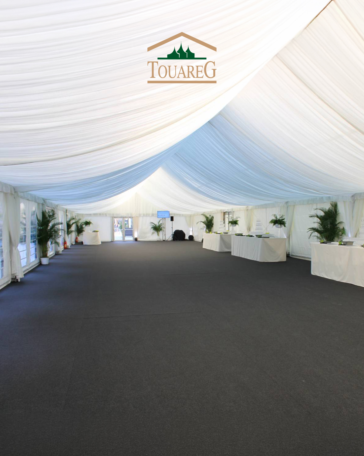 Intérieur d'une grande tente blanche pour événement, plafonds drapés, tables longues et plantes vertes; logo Touareg au centre.