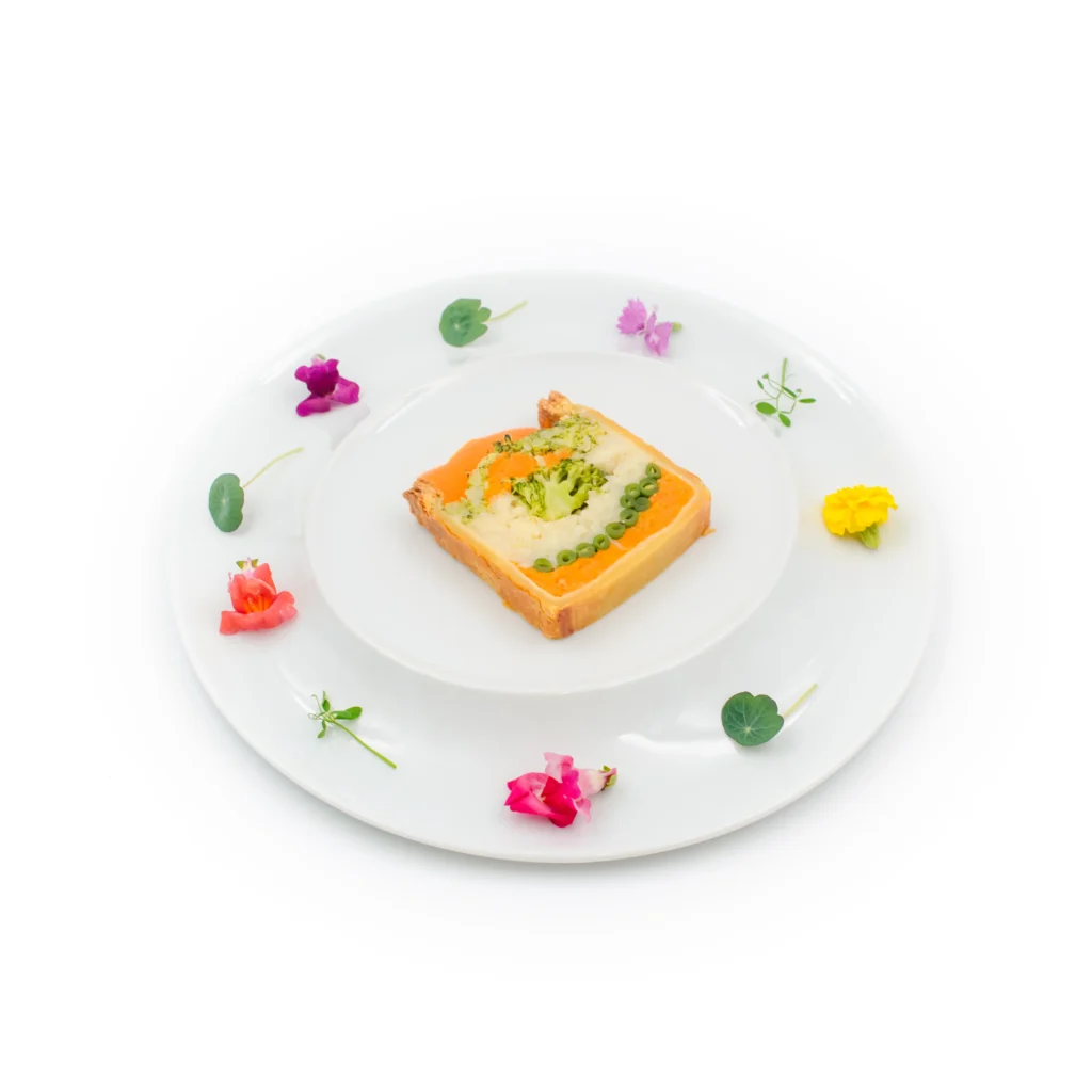 Tranche de pain garnie d'un coulis orange et herbes, décorée de fleurs comestibles sur assiette blanche.